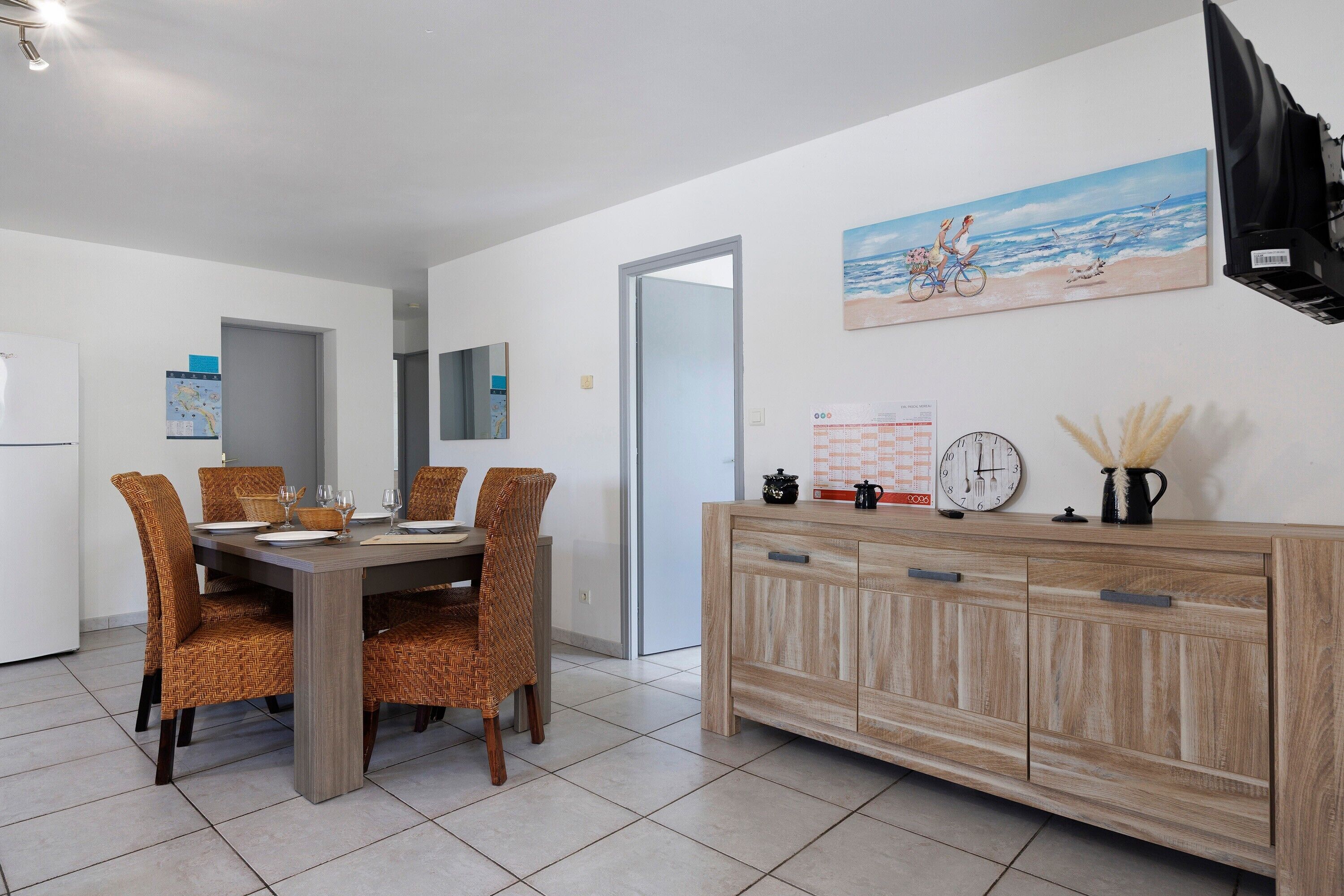 Ein Essbereich mit Holztisch und Korbstühlen, einem Holzkasten mit Schubladen, einem an der Wand montierten Fernseher und einem Gemälde mit Strandmotiv.
