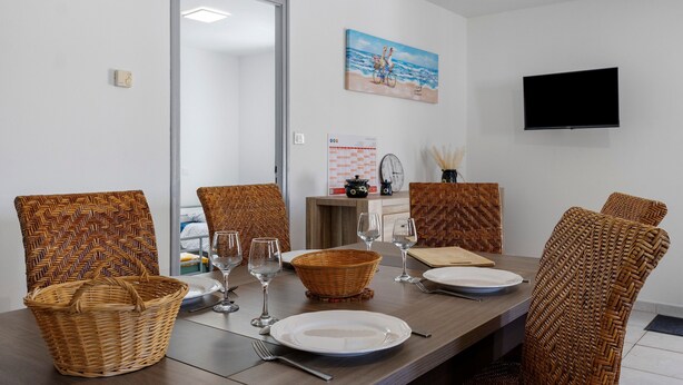 Uma sala de jantar com cadeiras de vime, uma mesa de madeira e uma televisão fixada na parede.