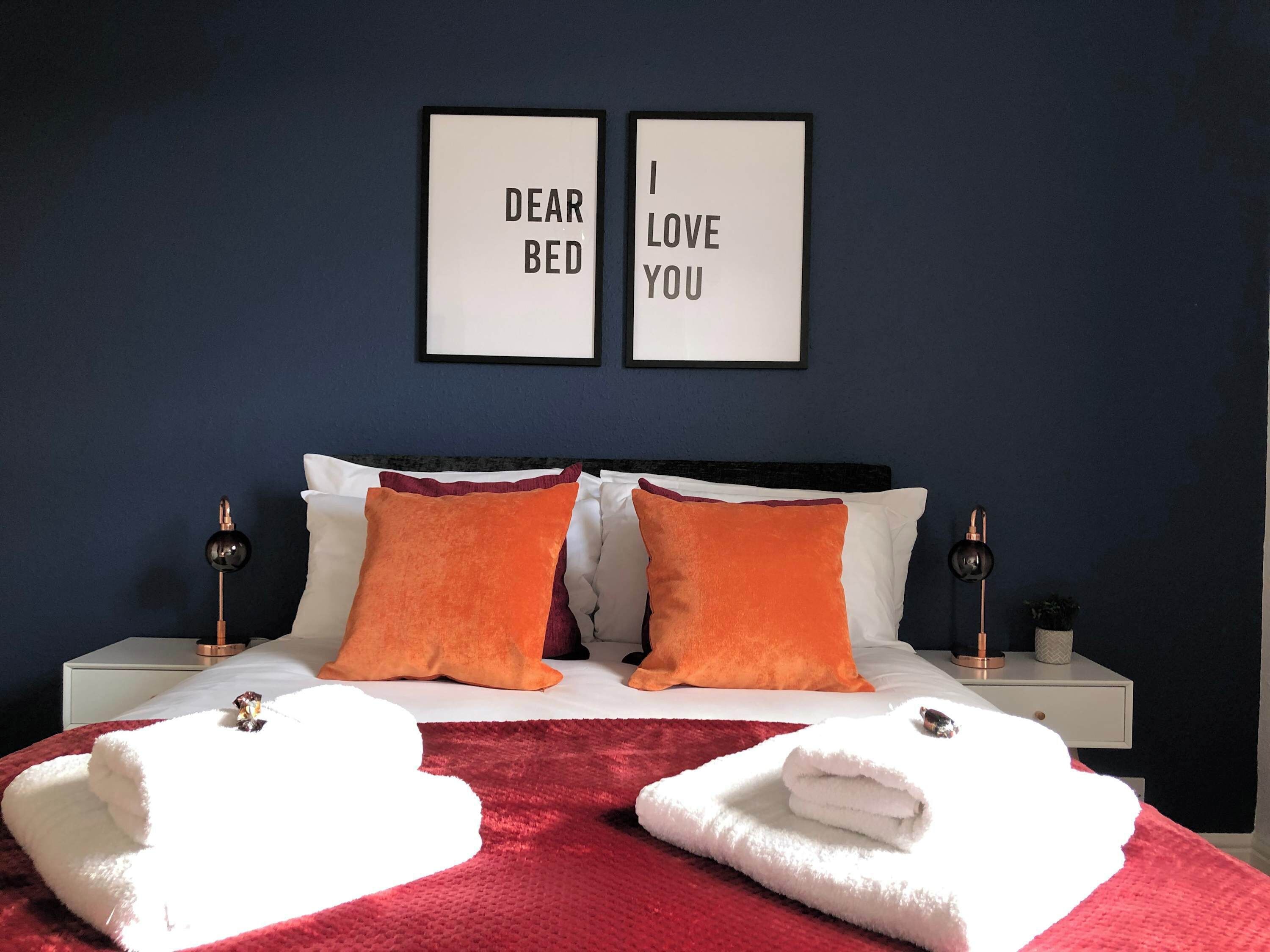 Un dormitorio con una cama, dos almohadas, dos mesitas de noche, dos cuadros enmarcados y dos toallas.