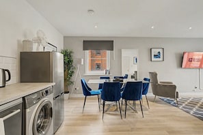 A modern kitchen with a washing machine, a dining area with blue chairs, and a view of a brick building through the window. - GuestReady - Chic 2bed with Balcony in Liverpool (Liverpool)