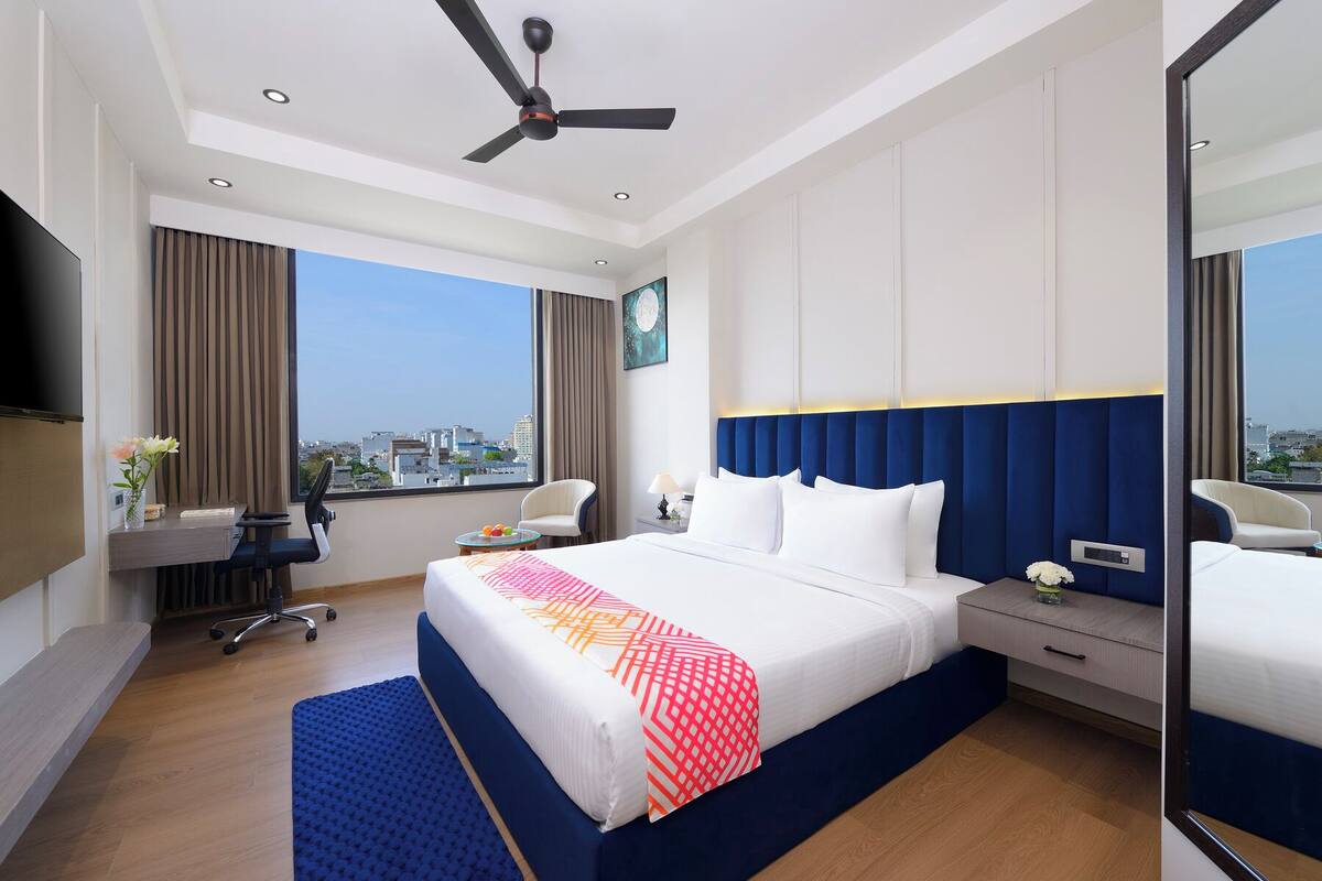 A hotel room with a large bed, a desk, a chair, a desk lamp, and a view of the city.
