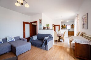 Ein Wohnzimmer mit einer Sofagarnitur, einem Esstisch mit Stühlen, einer Küchenzeile und Holzdielen.