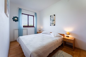 Ein Schlafzimmer mit Bett, Nachttischen, einem Fenster mit Vorhängen, einer Heizung und einem Wandbild.