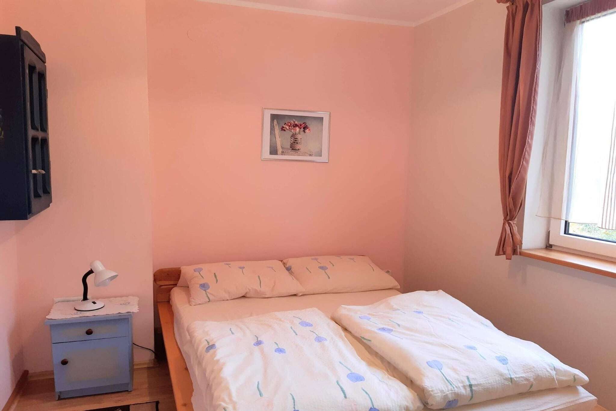 A bedroom with a bed, a bedside table with a lamp, a small framed picture on the wall, and a window with curtains.