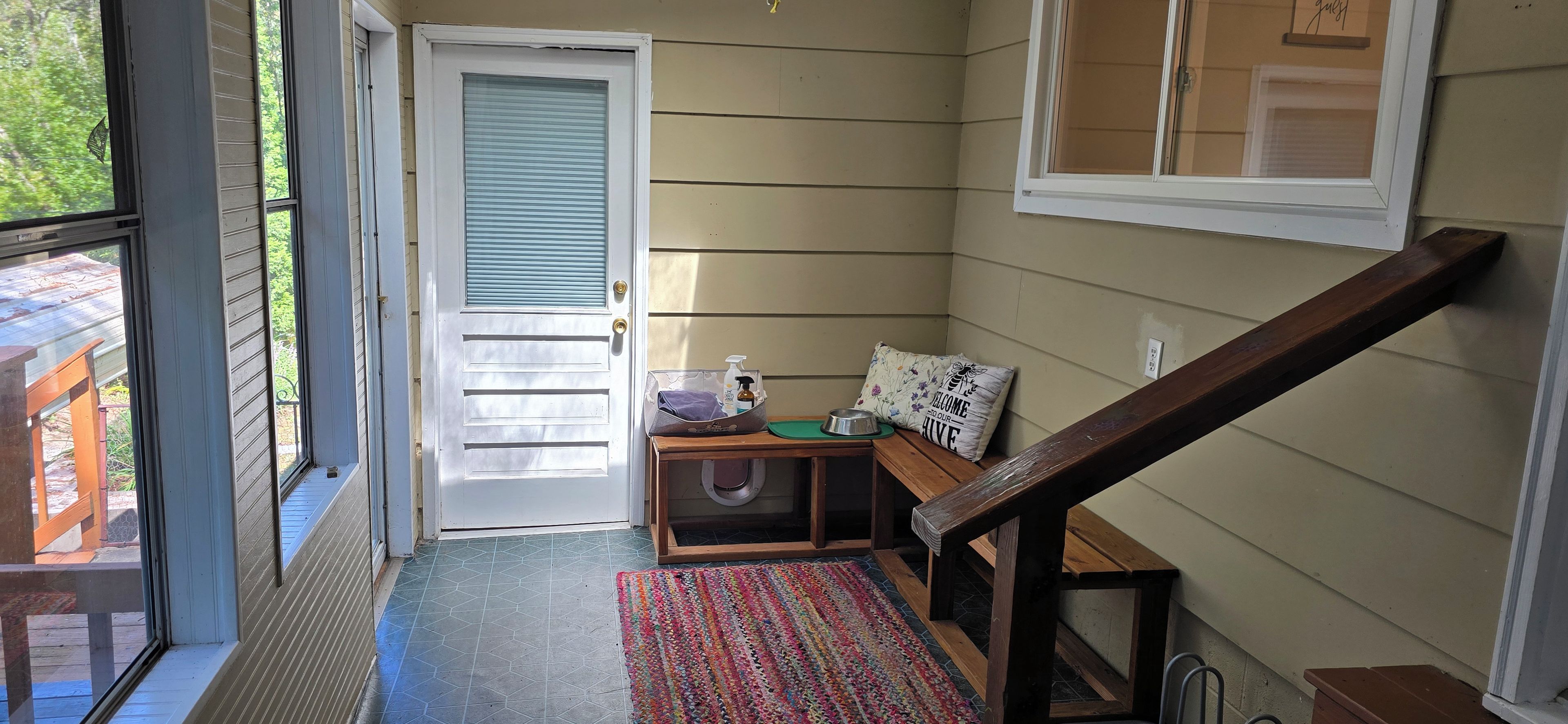 Ein Zimmer mit Holztür, einem kleinen Tisch mit Schale und Flasche, einem farbenfrohen Teppich und einem Fenster mit Jalousien.