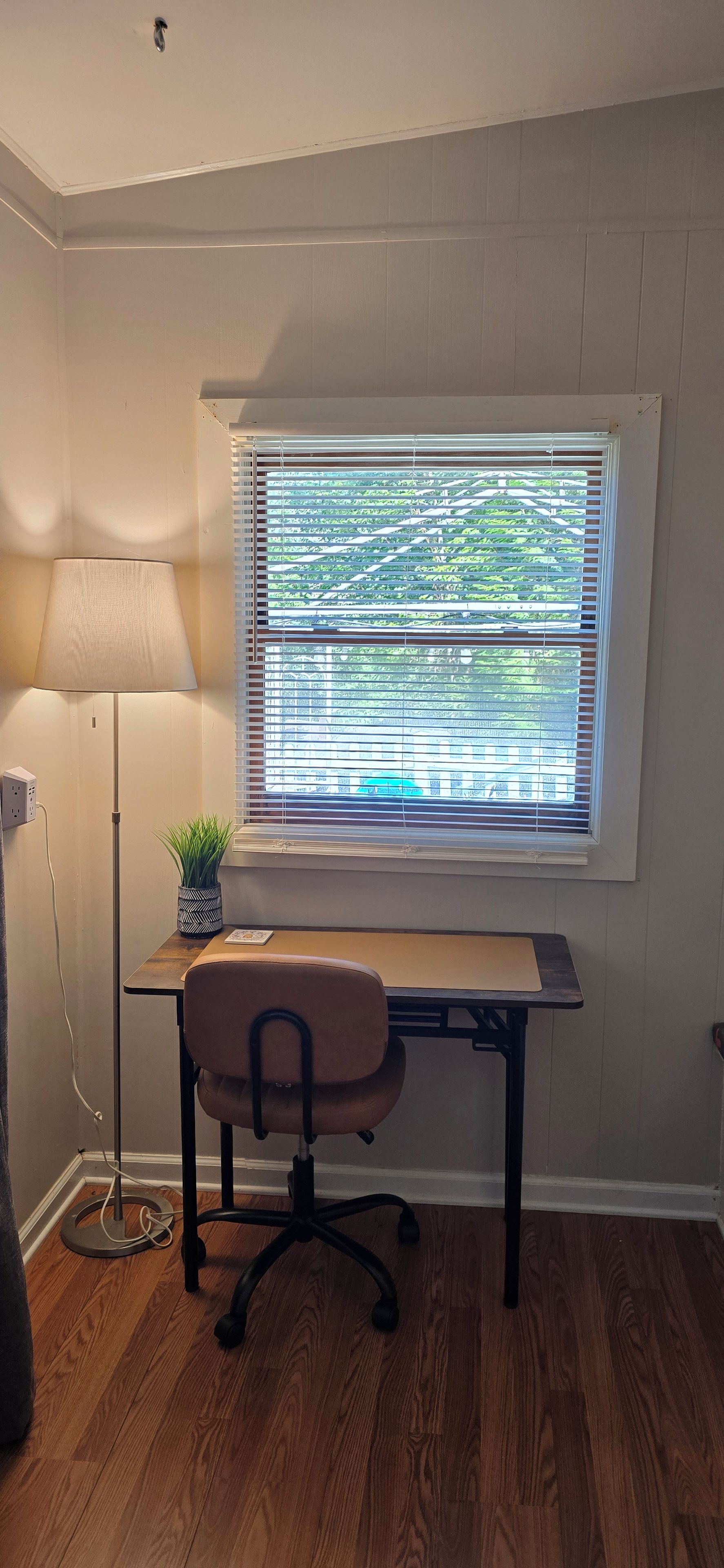 Une pièce comprenant un bureau, une chaise, une lampe et une fenêtre avec des stores.