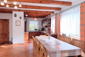 A dining area with a long table, wooden chairs, a vase with flowers, and a kitchen in the background.