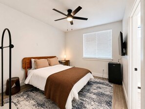 A bedroom with a bed, a ceiling fan, a TV, a window with blinds, and a small table with a lamp. - Bishop Arts 6BD/6BA Townhouse, Parking, Sleeps 12 (Dallas)