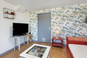 A room with a television on a white stand, a framed picture on a coffee table, a red bed, and a grey door. - Gîte at Pierrot and Pierrette’s, near Honfleur (La Rivière-Saint-Sauveur)