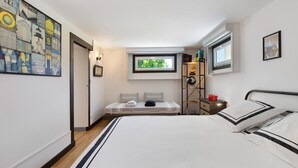 Un dormitori amb un llit, una tauleta de nit, una estant amb llibres, una finestra i una paret decorada amb obres d'art.