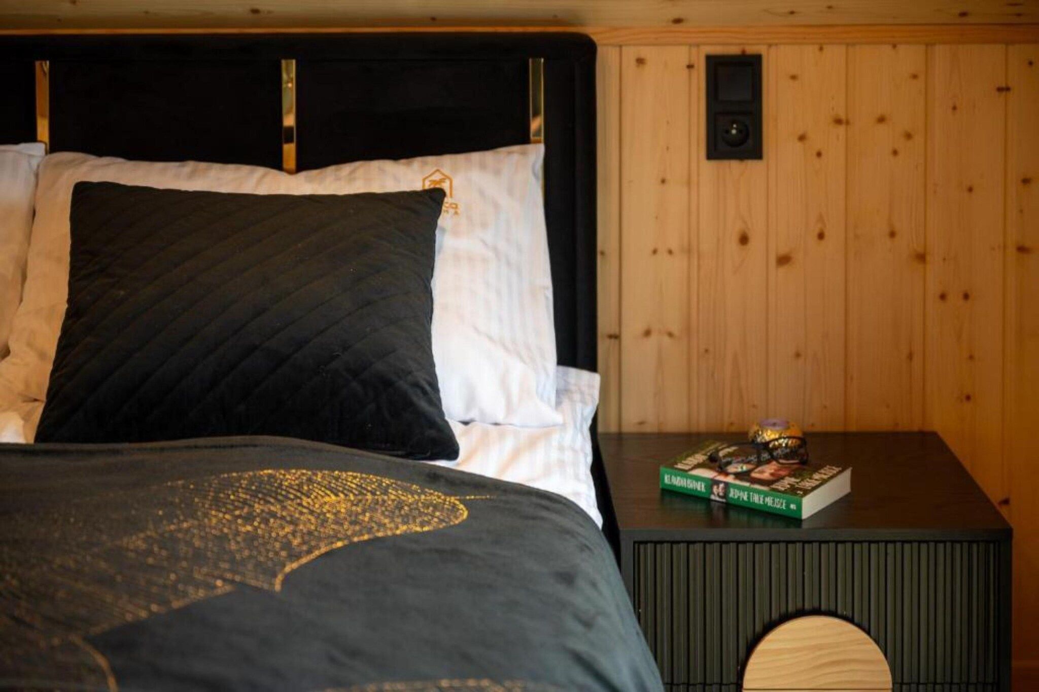 Un lit avec un oreiller noir et blanc, un livre sur une table de chevet et un mur en bois.