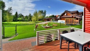 Ein überdachter Außenbereich mit Tisch und Stühlen, einer Rasenfläche und einem Holzgebäude im Hintergrund.