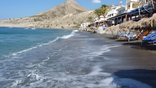 A beach with a mountain in the background, a clear sky, and a building with a thatched roof. - Apartment 'Maria Suite 2' with Sea View, Wi-Fi and Air Conditioning (Mirtos)