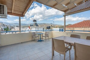 Ein überdachter Außenbereich mit Tisch und Stühlen, ein blauer Tisch und Blick auf Gebäude und Berge.