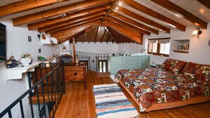 A loft bedroom with a wooden ceiling, a bed with a patterned bedspread, a desk with a vase of flowers, and a staircase.