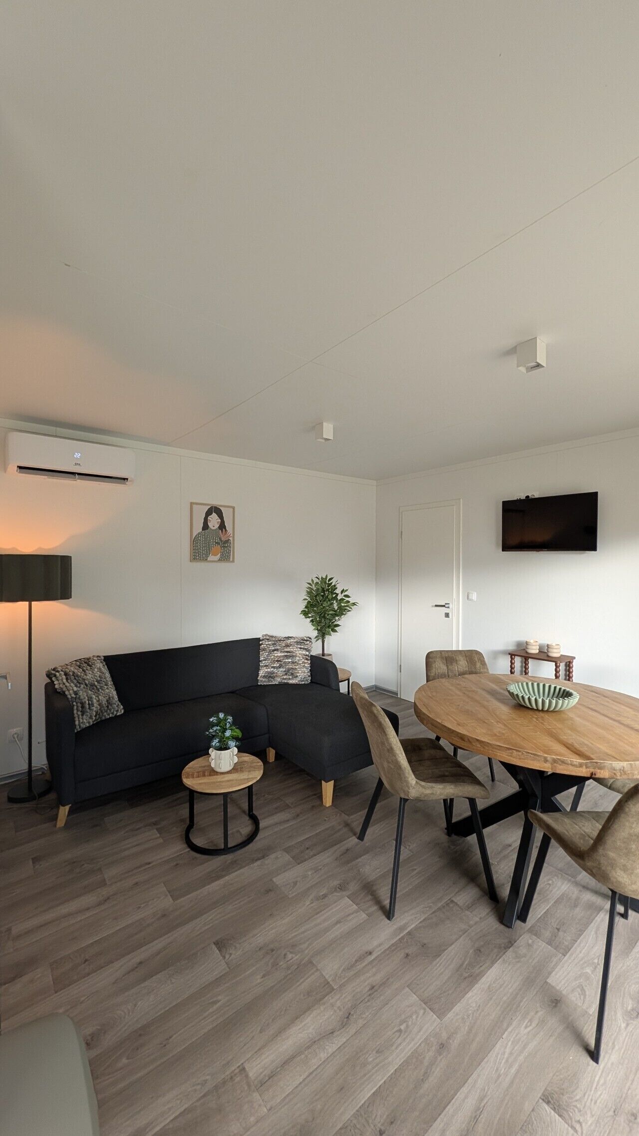 Una sala moderna con un sofá negro, una mesa de centro redonda, una mesa de comedor de madera con sillas, un televisor de pantalla plana montado en la pared y una planta en maceta.