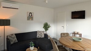 Un soggiorno moderno con un divano nero, un tavolino rotondo, un tavolo da pranzo in legno con sedie, una TV a schermo piatto montata a parete e una pianta in vaso.