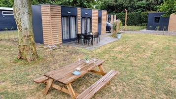 A wooden picnic table with a blue plate and wine glasses, set in a grassy area with a modern building in the background.