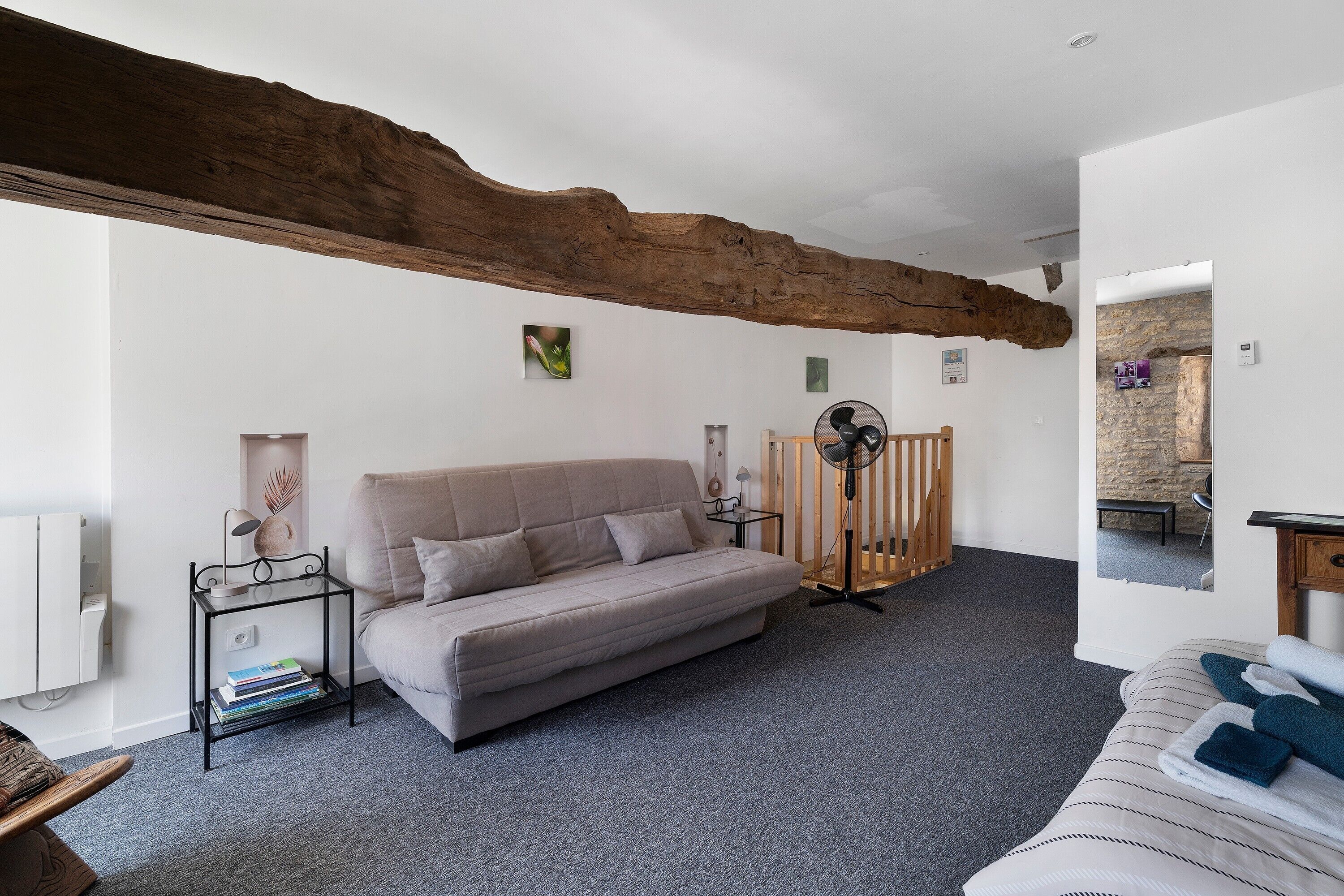 Une pièce comprenant un canapé, une petite table d’appoint avec des livres, un plafond avec une poutre en bois, un miroir et une vue sur un espace extérieur.