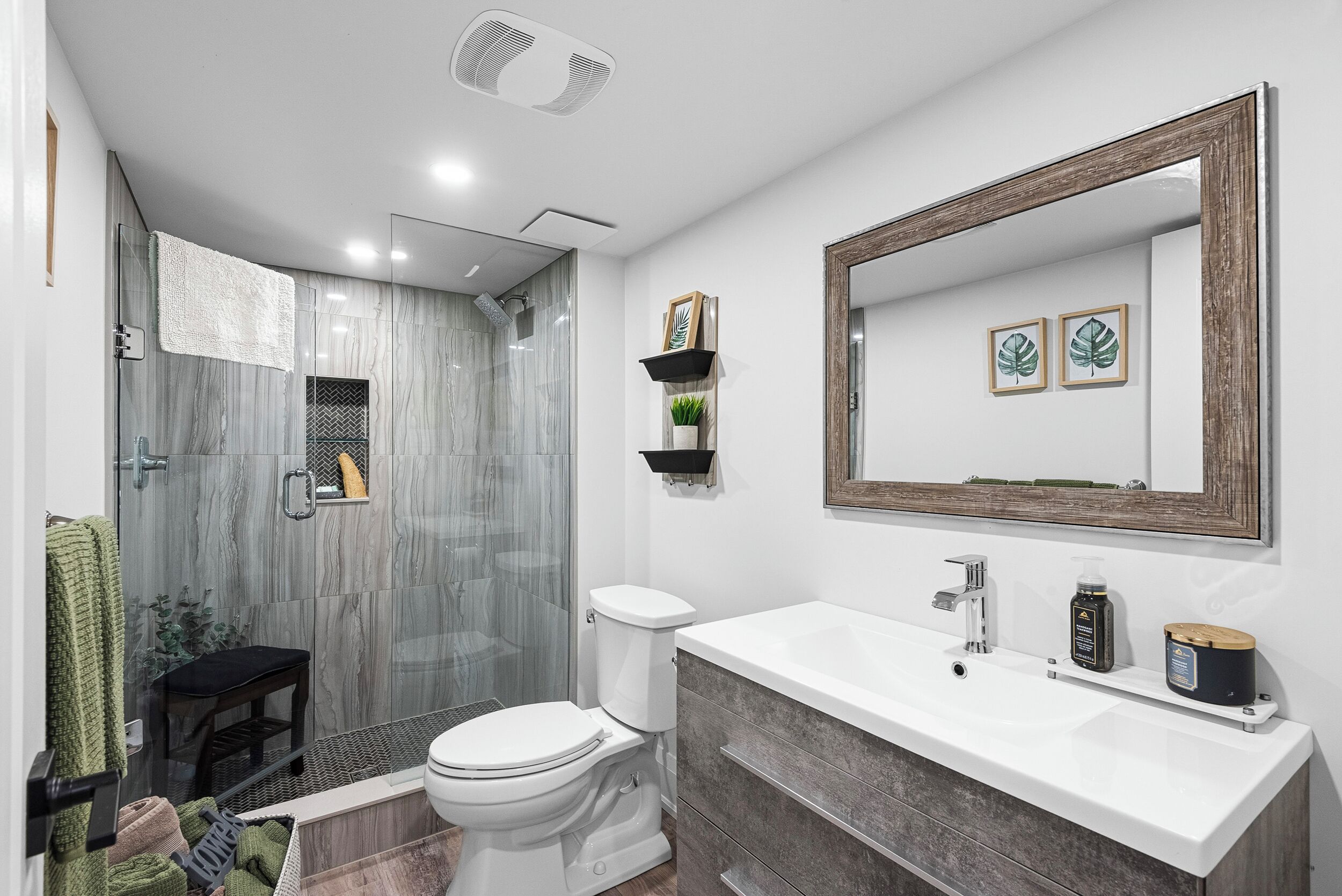 A modern bathroom with a glass shower enclosure, a white toilet, a large vanity with a mirror, and a small shelf with decorative items.