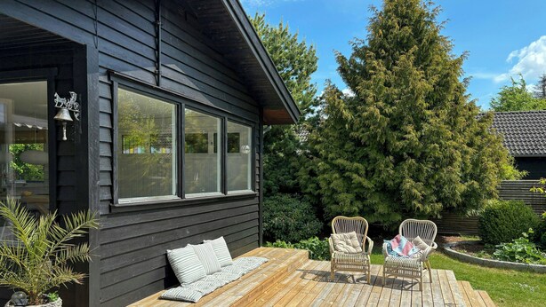 Una terraza de madera con dos sillas de mimbre y un banco, un árbol grande y una casa con revestimiento oscuro y ventanas amplias.
