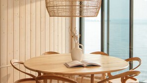A round wooden table with six chairs, a woven hanging light fixture, and a view of the sea through large windows. - Away Cabins (Roseneath)
