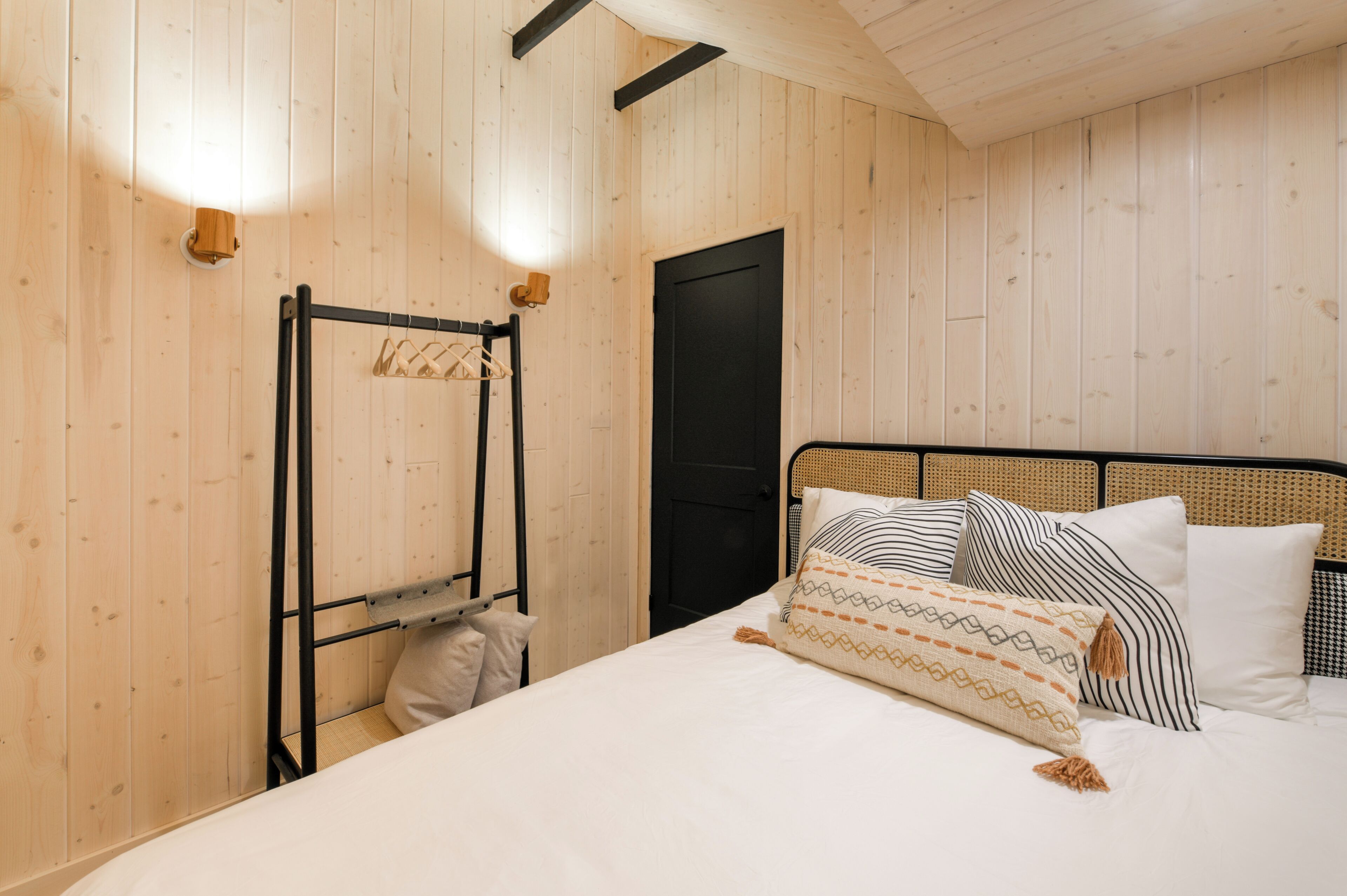 Een kamer met houten lambrisering, een bed, een zwarte metalen kapstok en een zwarte deur.