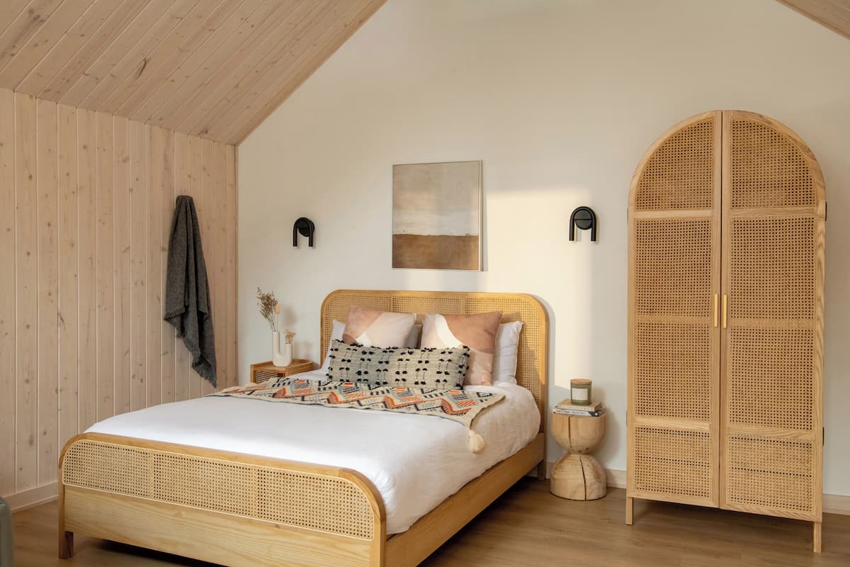 A room with a wooden bed, a rattan wardrobe, a wall-mounted artwork, and a small side table with a vase.