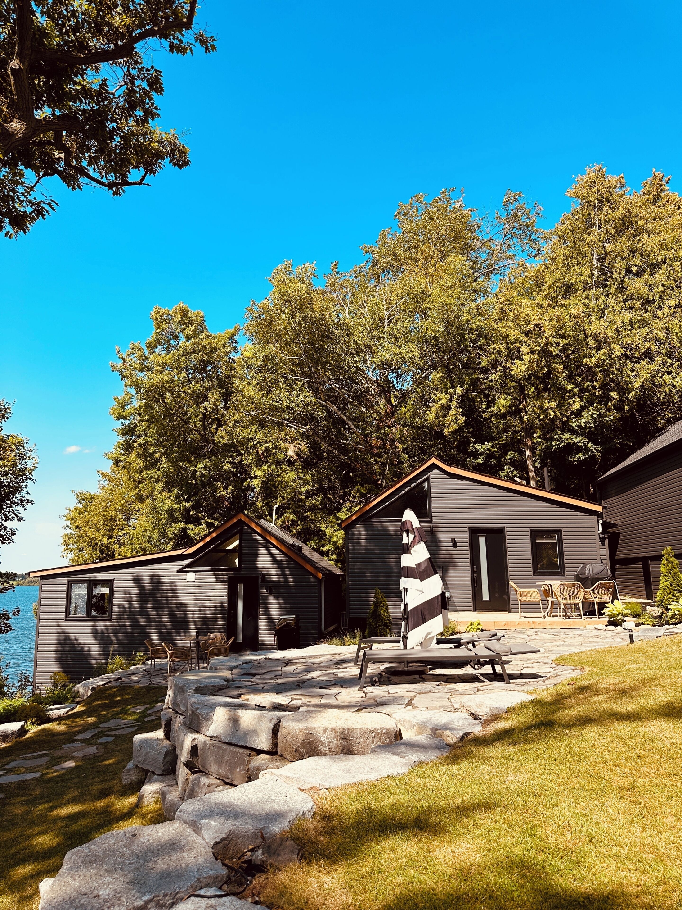 A lakeside cabin with stone steps leading to a dock, surrounded by trees and a clear blue sky.