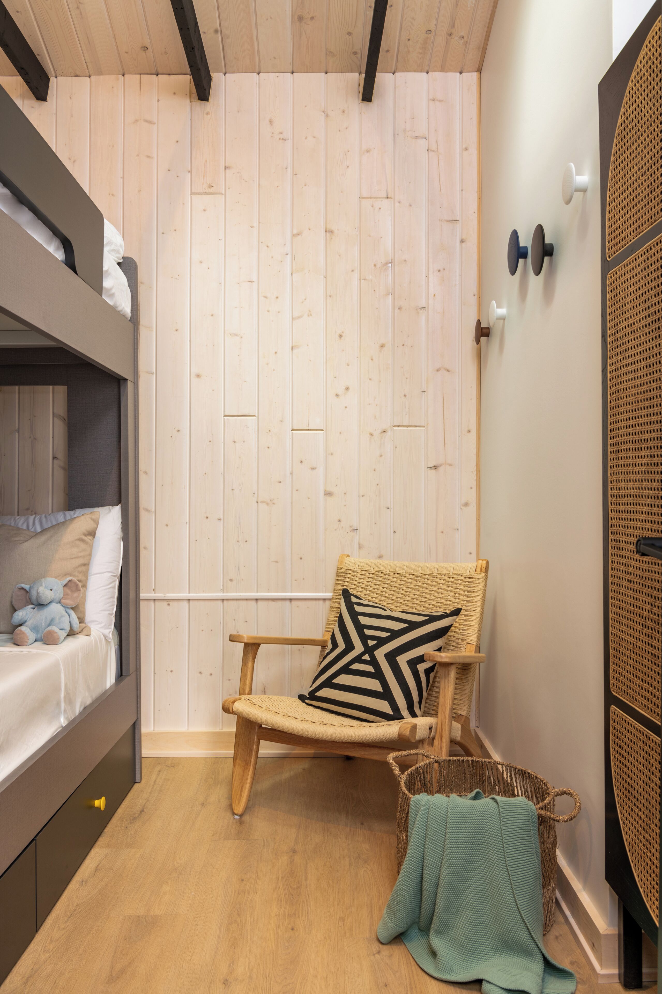 Een kamer met een stapelbed, een rieten stoel met een geometrisch kussen, een geweven mand en een turquoise plaid op de vloer.