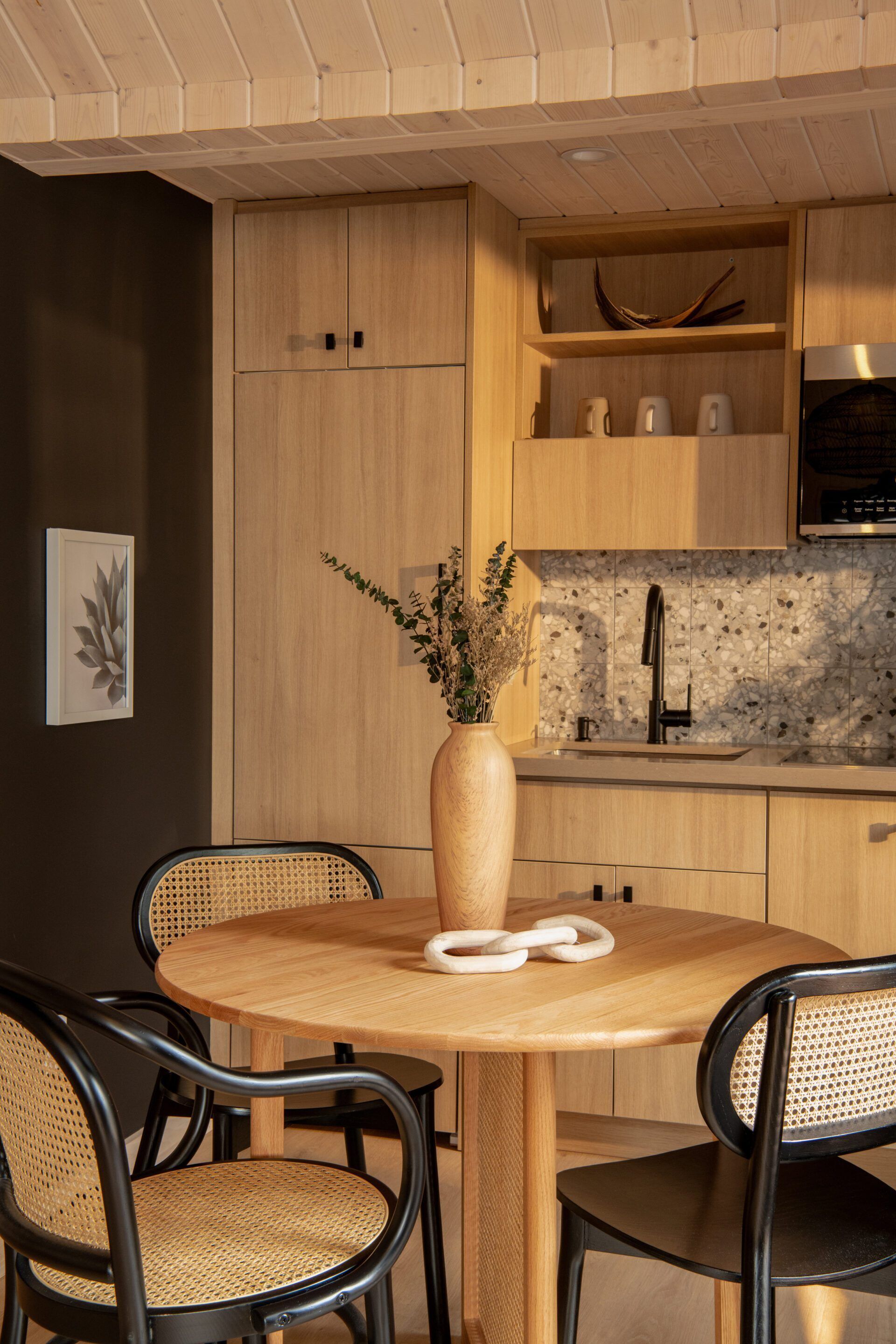 A modern kitchen with wooden cabinets, a round dining table with chairs, and a vase with dried flowers.