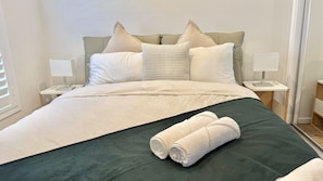 A neatly made bed with a green bedspread, white pillows, and two rolled towels. There are bedside tables with lamps and a window with blinds.