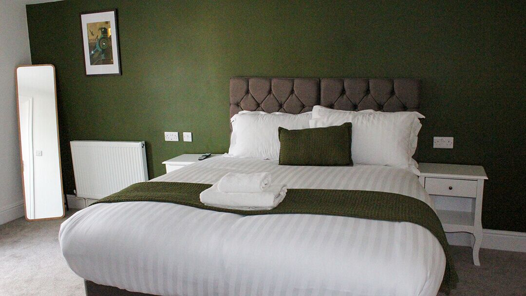 A neatly made bed with white linens and a green throw, a bedside table with a lamp, and a framed picture on the wall.