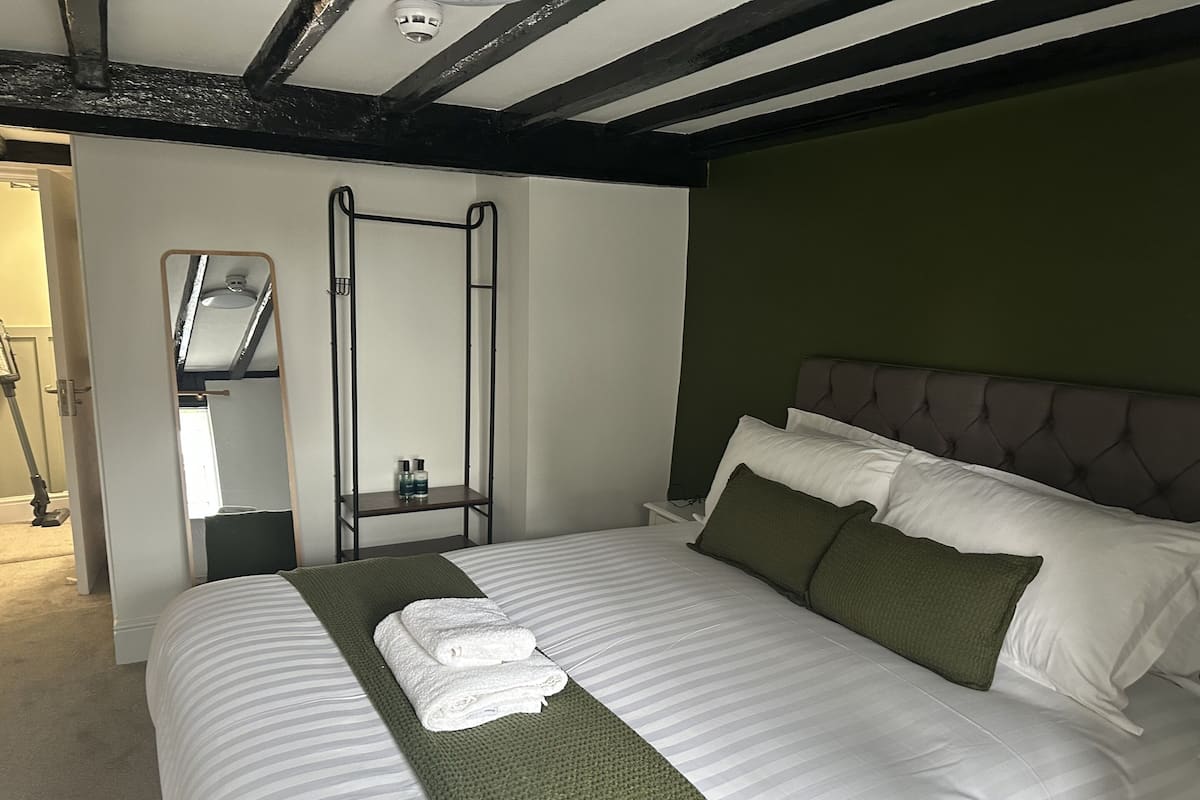 A neatly made bed with white linens and green accents, a bedside table with a bottle, and a wall-mounted ladder.