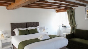 Quarto com cama, mesas de cabeceira, uma cadeira, janela com cortinas e teto de madeira.