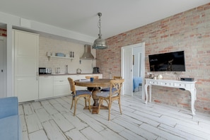 Une cuisine avec des meubles blancs, un mur en briques, une table à manger avec des chaises, un téléviseur posé sur un meuble blanc et un luminaire au plafond.