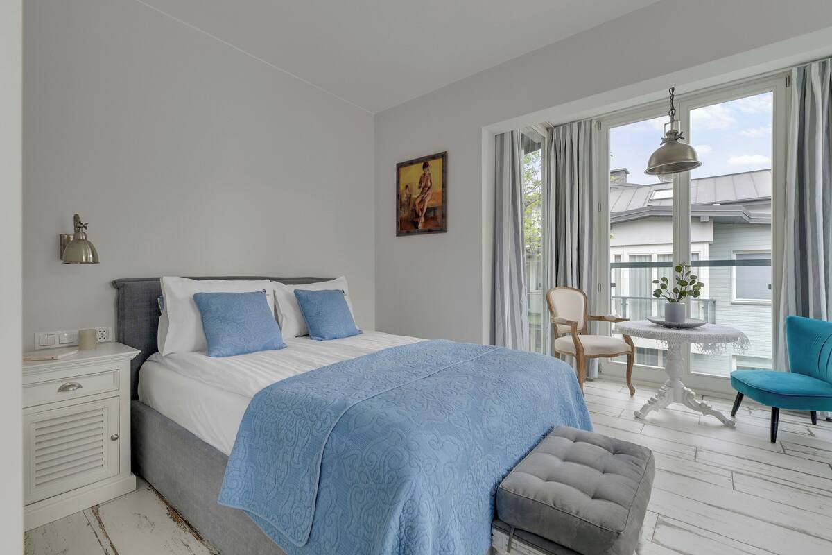 Un dormitorio con cama, mesita de noche, lámpara, silla y vistas al exterior.