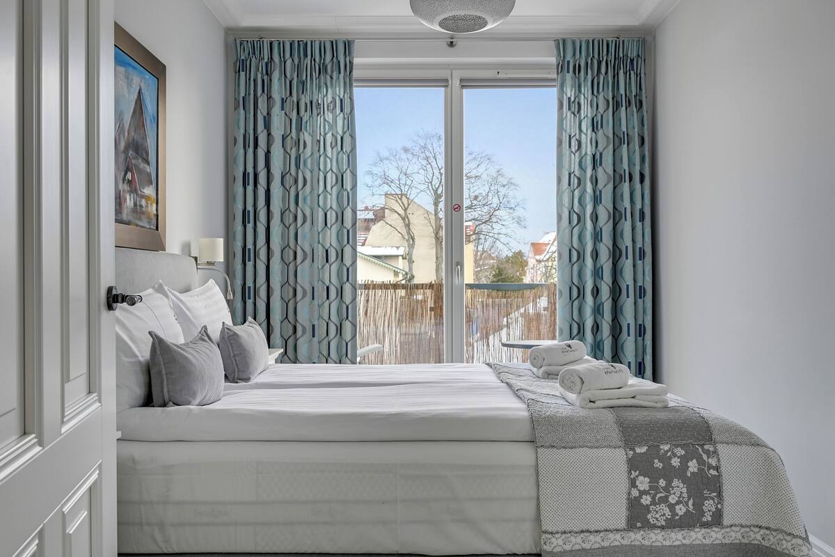 Habitación de hotel con cama, un cuadro, ventana con cortinas y vistas a edificios.