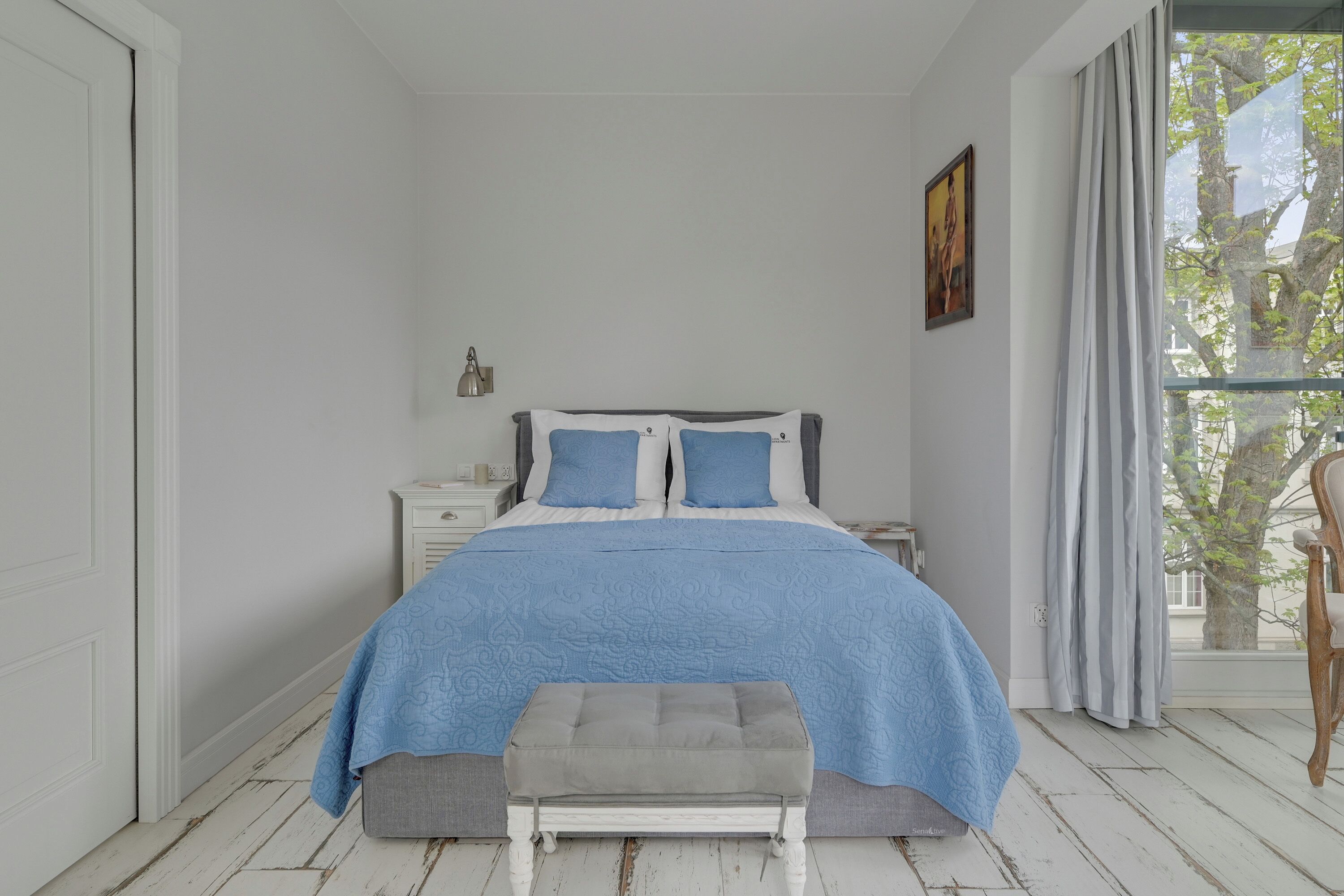 A bedroom with a bed, a bench, a nightstand, a lamp, and a window with curtains.