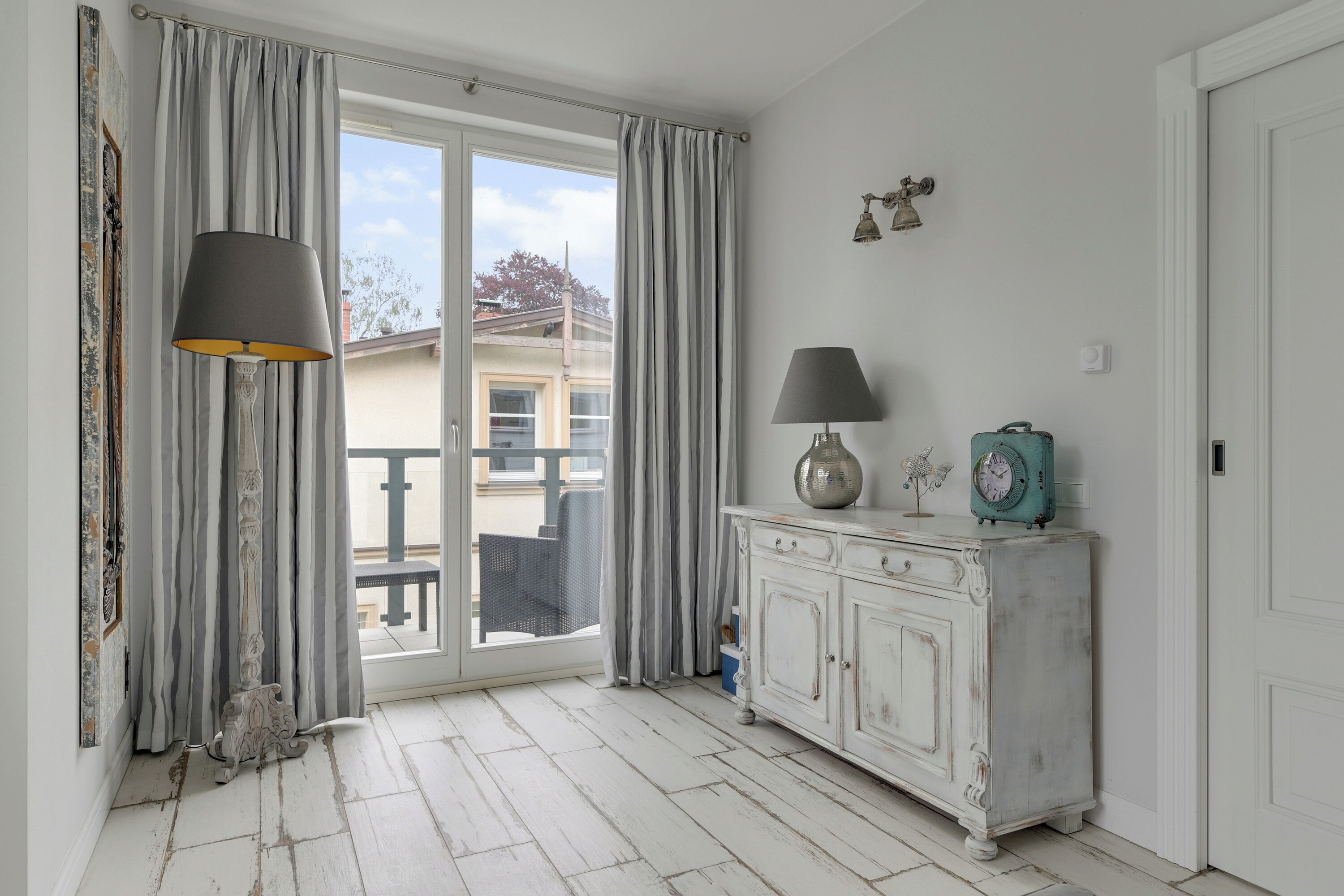 Una habitación con piso de madera blanca, un armario blanco con un reloj y un ventanal corredero a un balcón exterior.