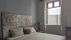 Una cama bien hecha con un cabecero decorativo, una lámpara de noche y una ventana con una persiana abierta.