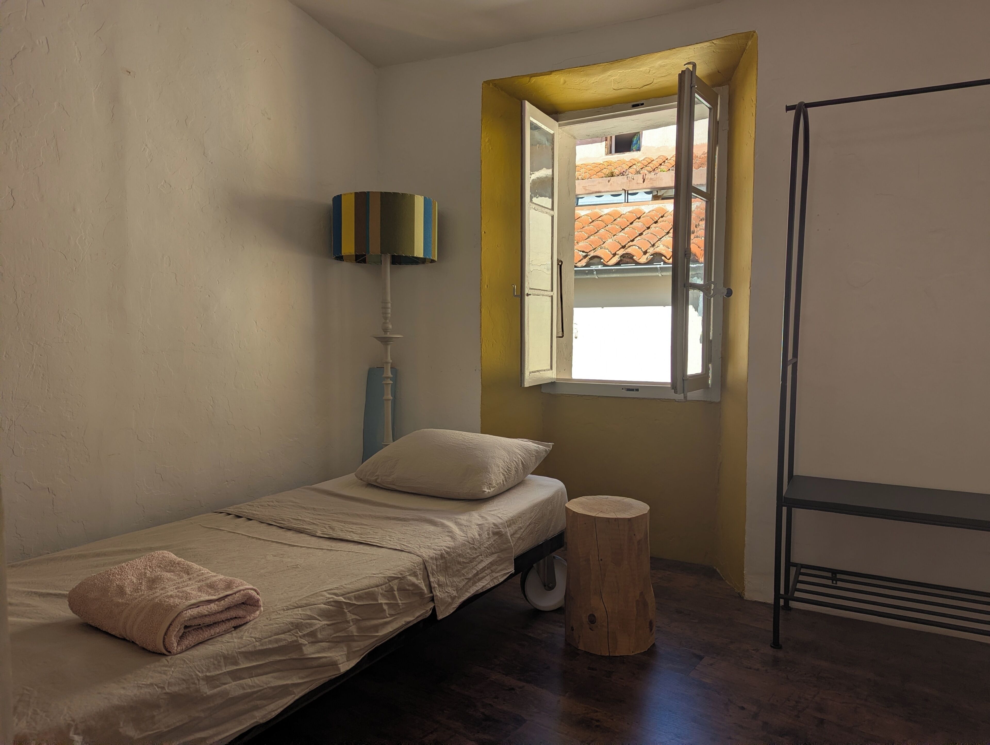 Une petite chambre avec un lit simple, un tabouret en bois, une lampe sur pied et une fenêtre dont le volet est ouvert.