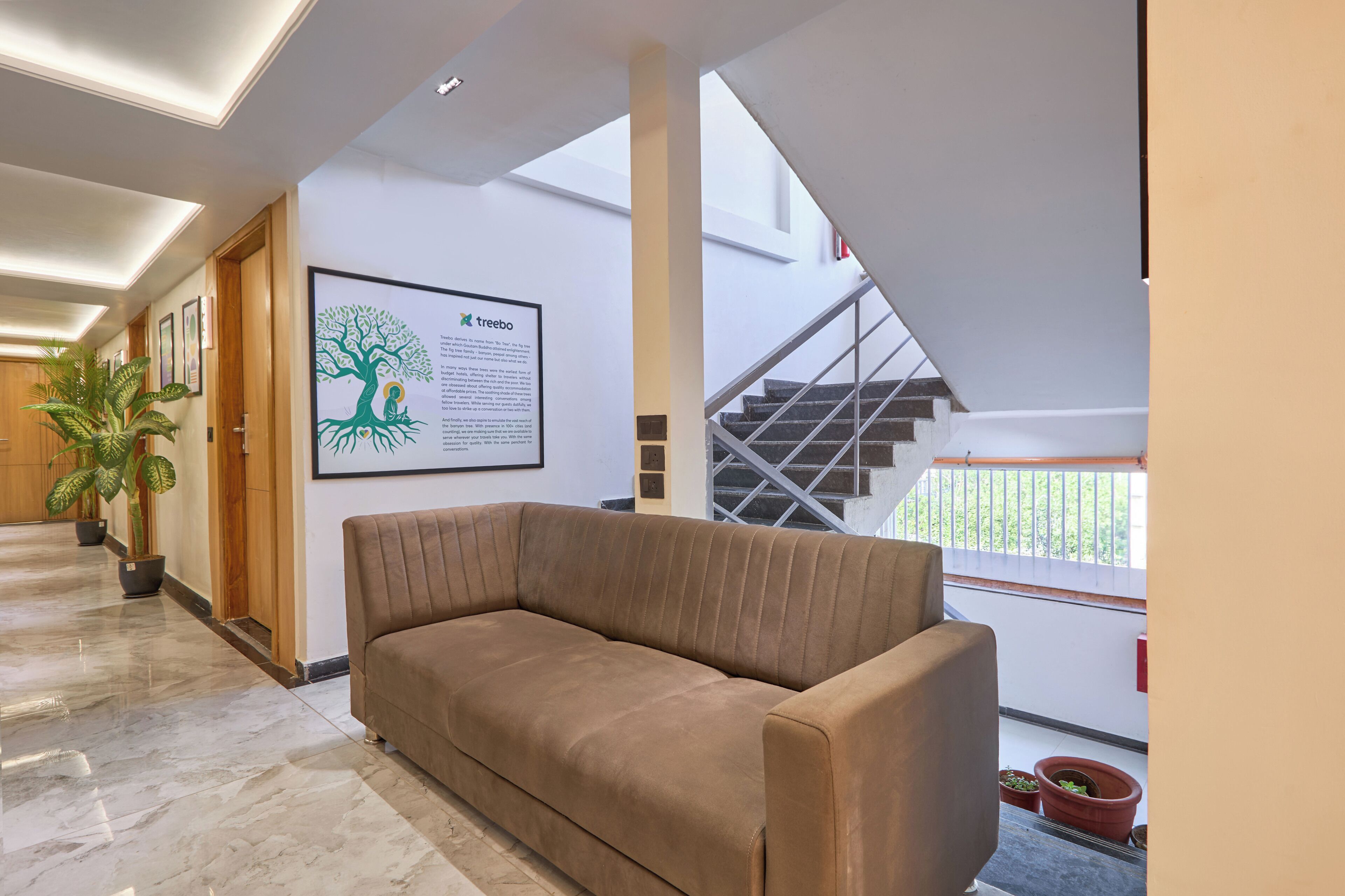 A modern interior with a brown sofa, a framed informational poster, potted plants, and a staircase leading to a balcony.