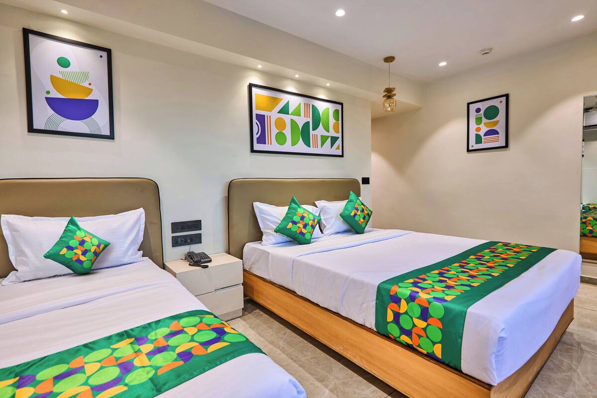 A hotel room with two beds, each with a green patterned bedspread, white bedding, and green pillows. There are framed abstract artworks on the walls, a bedside table with a phone, and a mirror.