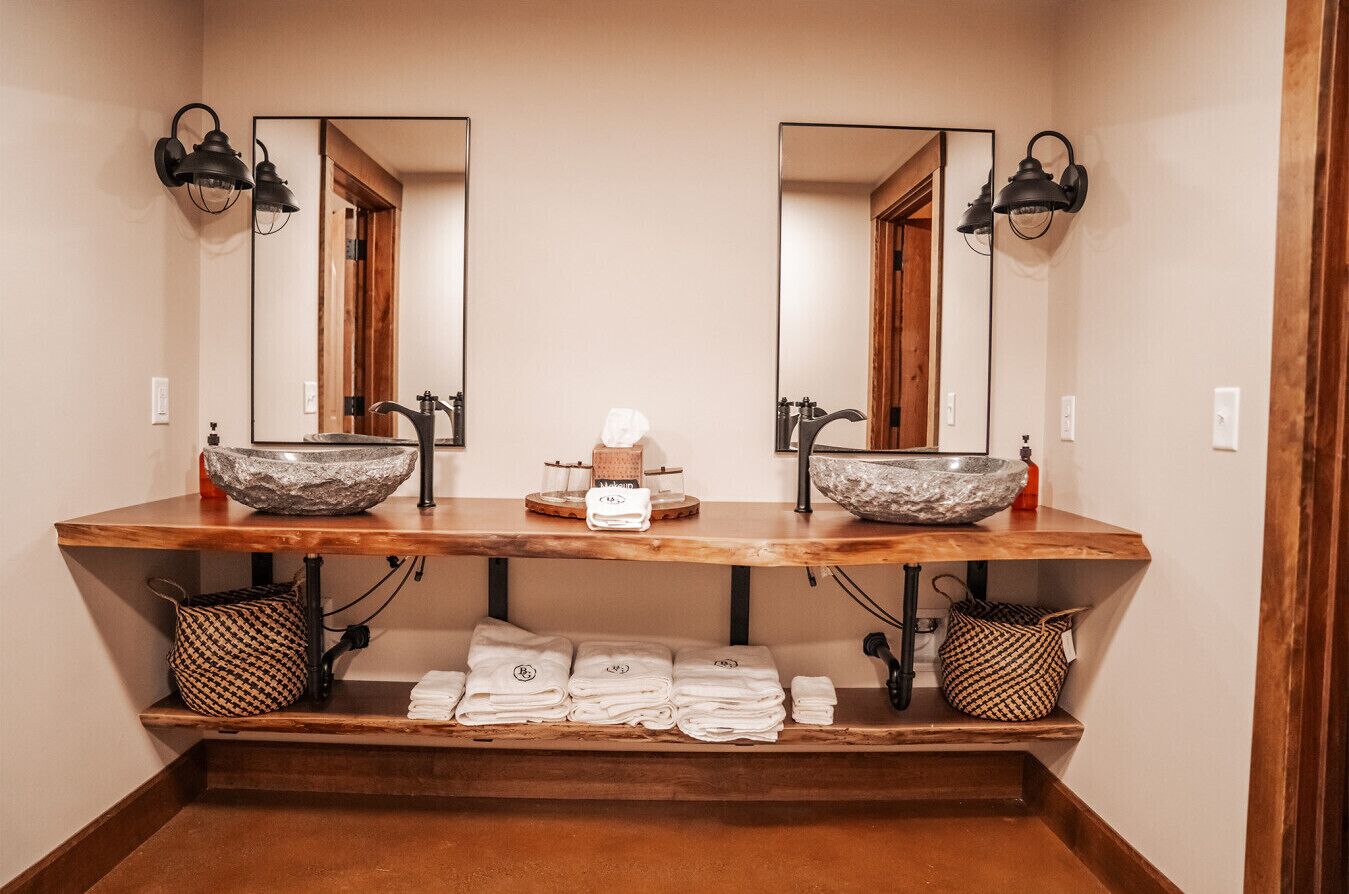 A bathroom with two sinks, each with a large stone basin and a mirror above. There are towels and a soap dispenser on the counter.