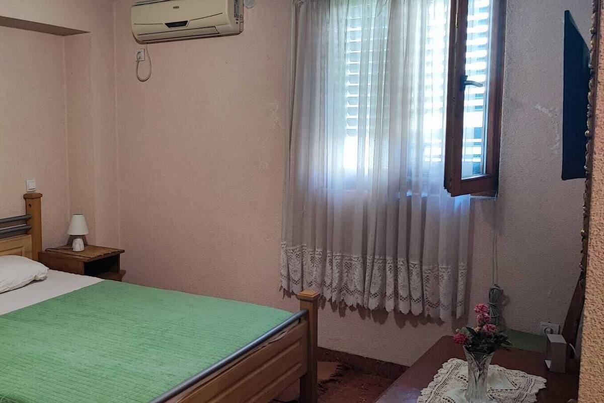A bedroom with a bed, two bedside tables, a wall-mounted air conditioner, a painting, and a window with curtains.