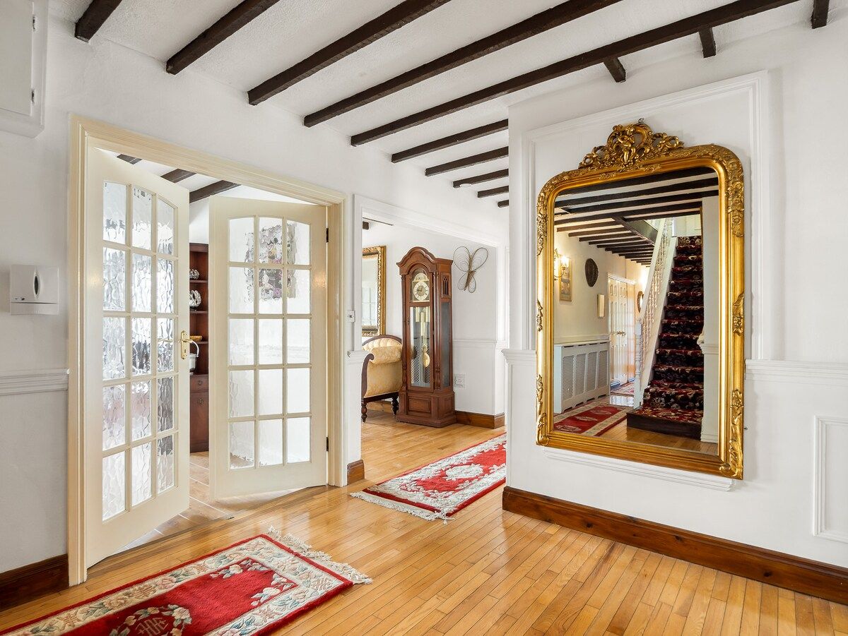 An interior with a wooden floor, a red and white patterned rug, a large ornate mirror, and a staircase with a red carpet.