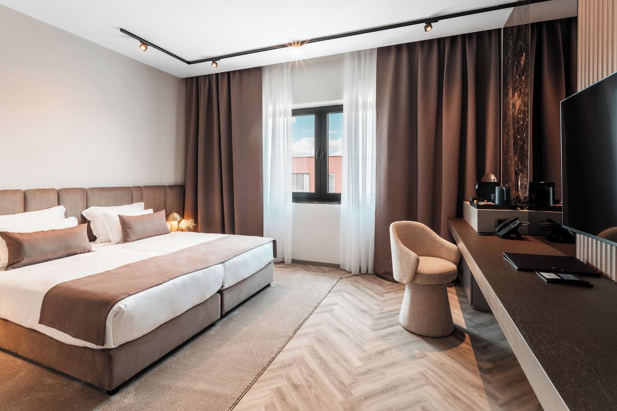 A modern hotel room with a large bed, a desk with a chair, a television, and a window with curtains.