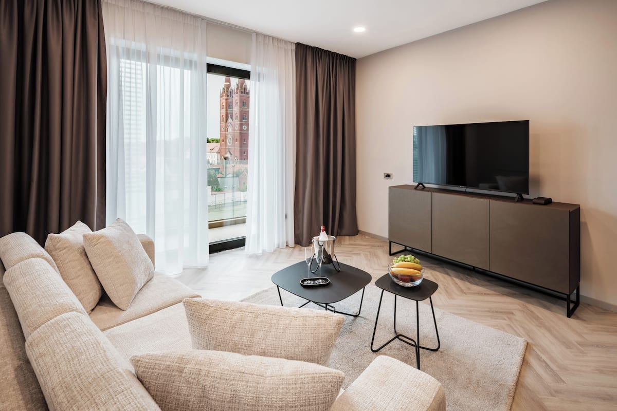 A modern living room with a sofa, a coffee table, a TV on a cabinet, and a view of a building outside.