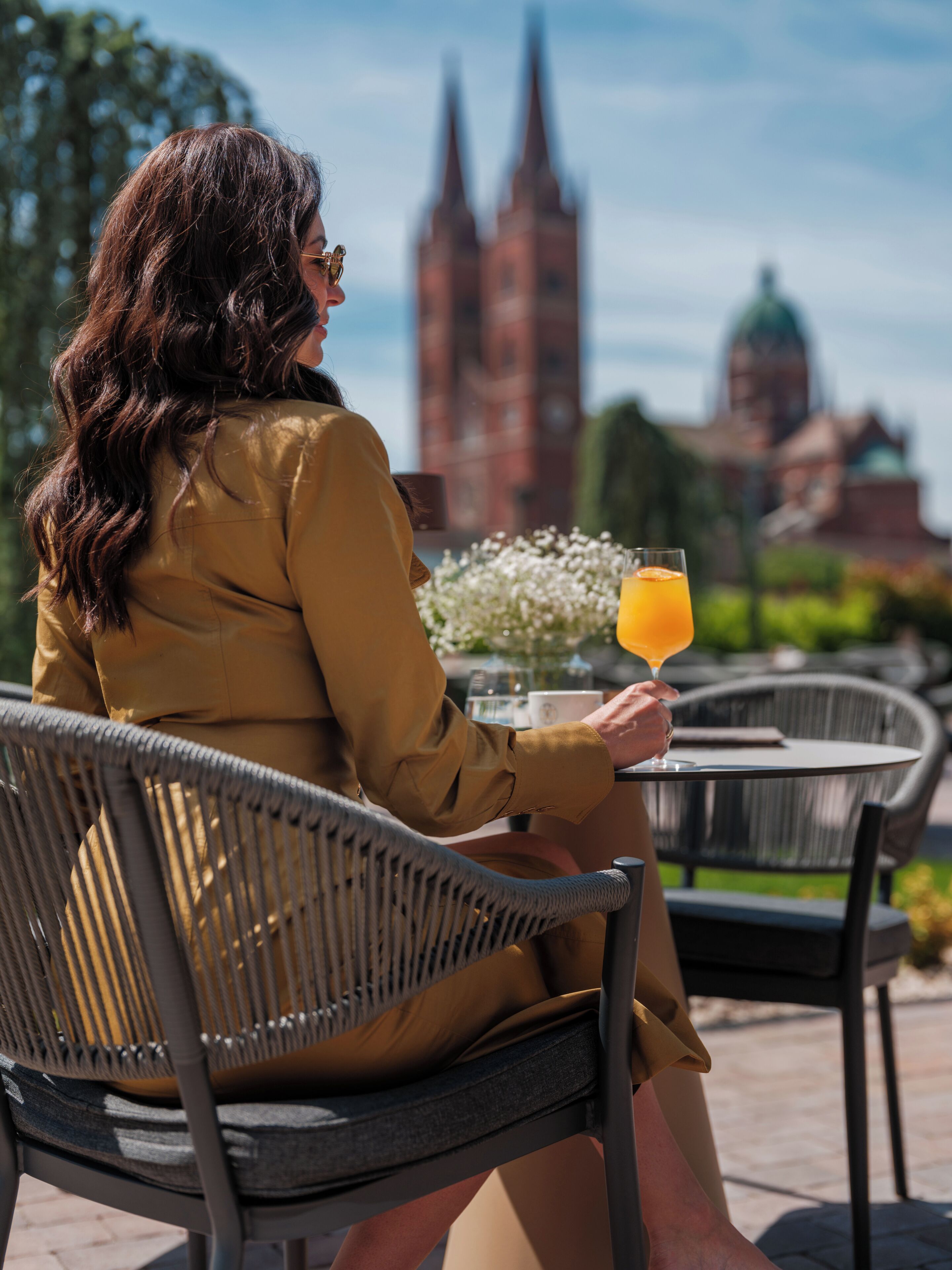 A person sitting at an outdoor table with a drink, overlooking a cityscape with notable architecture.